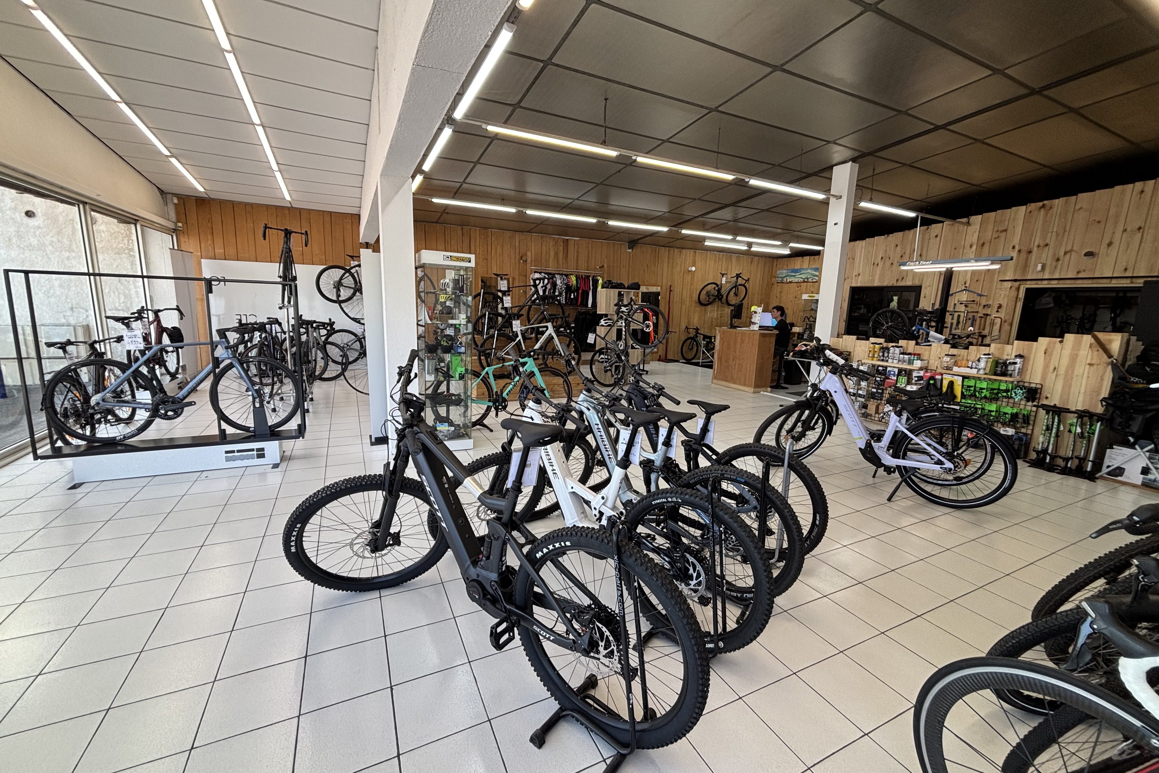Magasin de vélos intérieur avec plusieurs vélos exposés, y compris des modèles de montagne et électriques. Étagères avec équipements et accessoires en arrière-plan.